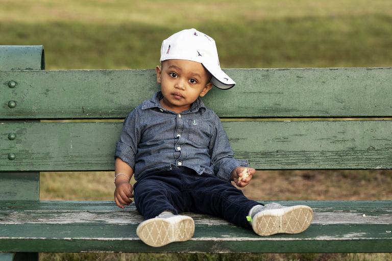 toddler boy sitting on green bench wearing hat blue shirt sneakers boston massachusetts herter park portrait